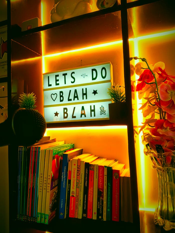 A bookshelf with neon lighting featuring a humorous sign, books, and decorative flowers.